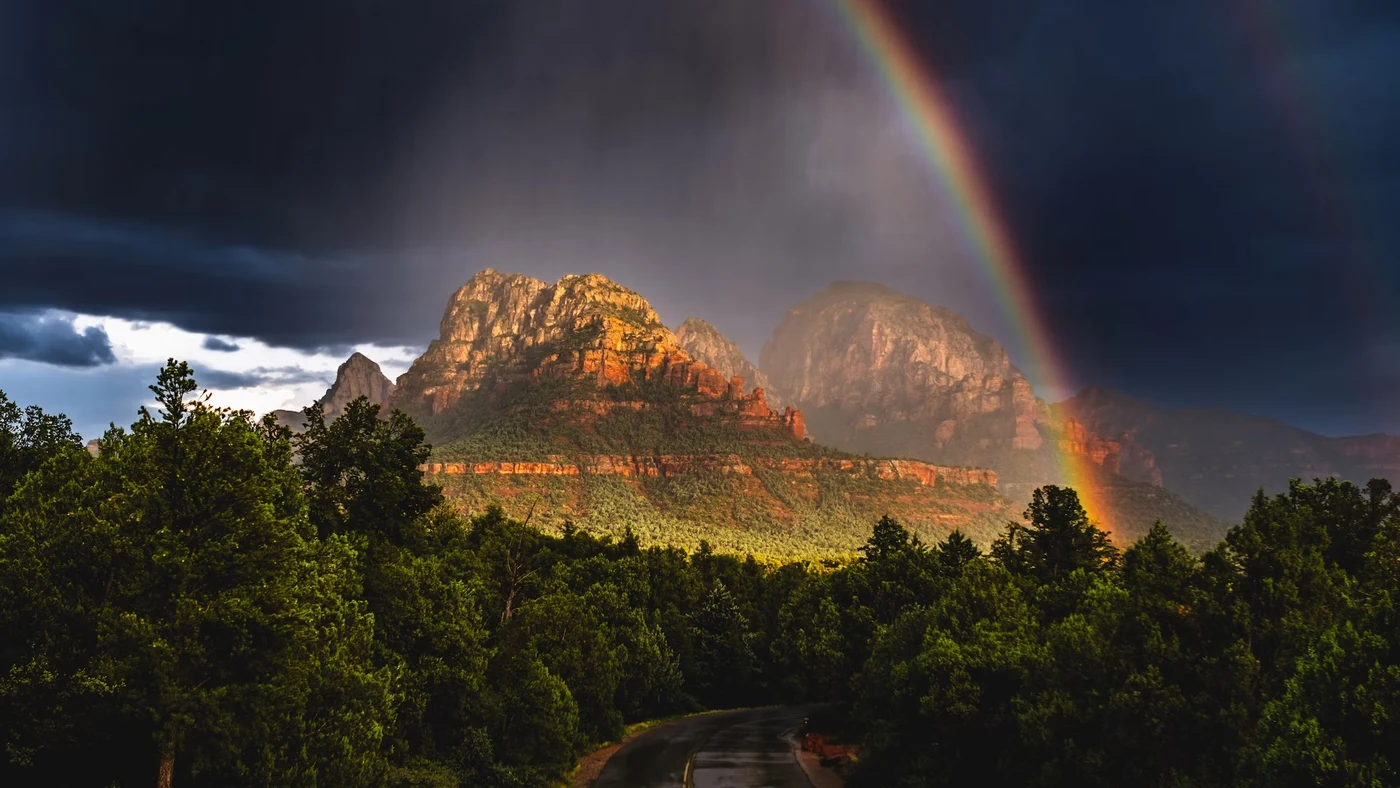 A double rainbow breaking through storm clouds over red rock mountains in Sedona, Arizona, after a monsoon thunderstorm.